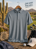 A slate blue-gray short-sleeve polo shirt on a wooden hanger, styled on a rustic table with golf clubs, a golf cap, a golf ball, and desert-themed props in the background. The text 'LOOK GOOD.